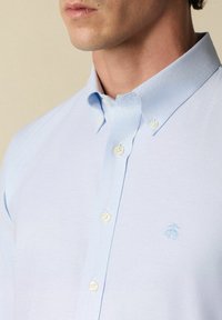 Lichtblauw overhemd met puntkraag, voorzien van witte knopen en een klein geborduurd logo op de borst. Gladde stofstructuur.