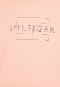 La scritta "HILFIGER" è contornata da linee rosse sottili con un bordo rettangolare doppio su uno sfondo di tessuto rosa chiaro.