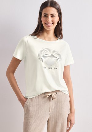 Femme souriante portant un T-shirt blanc à manches courtes avec un motif éclat de soleil et le texte « SUN SAND SEA », associé à un pantalon beige texturé avec cordon de serrage.