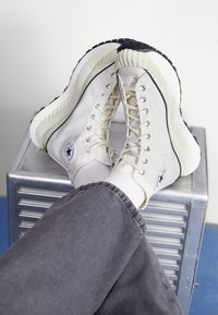 Beige hög topp sneakers med en texturerad gummisulor, rund logotypapplikation och silverfärgade öglor; i kombination med grå byxor och vita strumpor.