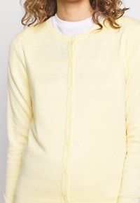Jasnożółty kardigan wykonany z miękkiego materiału, z okrągłym dekoltem, długimi rękawami oraz zapięciem na guziki. Gładka faktura z ryftowanymi akcentami.