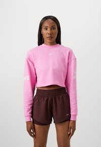 Różowy, krótki sweatshirt z długimi rękawami, z białym logo Nike na piersi i rękawie; zestawiony z ciemnobrązowymi szortami na elastycznej talii.