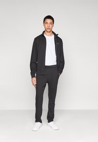 Γκρι αθλητικό μπουφάν με ψηλό γιακά και φερμουάρ μπροστά, συνδυασμένο με ταιριαστό παντελόνι στενής εφαρμογής και άσπρα αθλητικά παπούτσια. Λεπτό ύφασμα, μινιμαλιστικός σχεδιασμός.