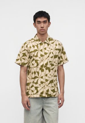 Jeune homme portant une chemise beige à manches courtes avec un grand motif floral vert et un jean bleu clair, debout devant un fond clair uni.