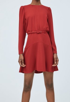 Rode jurk met lange mouwen, een getailleerde ceintuur, wijde rok en licht pofschouders, gedragen door een staande persoon tegen een effen achtergrond.