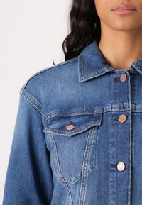Denim jacket in light blue, featuring a shirt collar, two chest pockets, and metallic buttons with a rose gold finish.