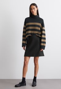 Maglione oversize a righe verde scuro con strisce beige, abbinato a una gonna nera. Modella indossa calzini e scarpe nere, in piedi su uno sfondo neutro.