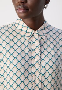 Button-down shirt featuring a geometric pattern in teal and beige on a cream background. Collared design with visible buttons.