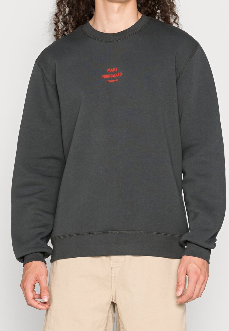 Personne portant un sweat-shirt gris foncé avec un petit texte rouge « Mads Nørgaard Copenhagen » sur la poitrine et un pantalon beige, debout devant un fond blanc.