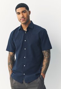 Camicia a maniche corte blu navy in tessuto liscio, con colletto button-down, bottoni bianchi e vestibilità comoda, indossata sopra pantaloni grigi.