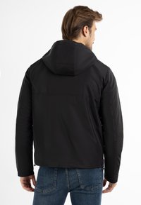 Uomo che indossa una giacca nera con cappuccio e jeans blu, di spalle contro uno sfondo bianco uniforme.