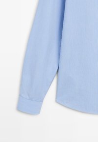 Lichtblauw overhemd met lange mouwen, dunne verticale witte strepen en een enkele knoopmanchet op een witte achtergrond.