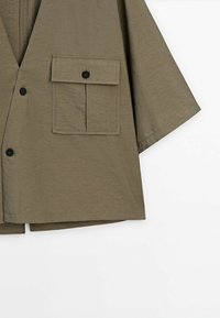 Chemise manches courtes vert olive avec boutons noirs, une grande poche à rabat sur la poitrine gauche et une coupe décontractée.