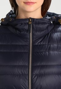 Dunkelblaue Steppjacke mit gequilteter Textur, silbernem Reißverschluss und verstellbarer Kapuze. Beinhaltet goldfarbene Hardware-Details am Kragen.