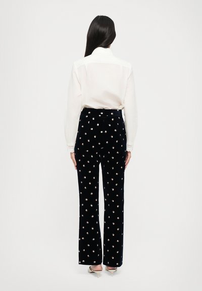 Tumši zili samta bikses ar izliektu dizainu, rotātas ar maziem ziedu rakstiem. Savienotas ar baltu garām piedurknēm krekli, skatoties no aizmugures.