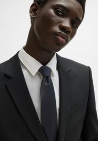 Costume noir à texture avec une poche poitrine simple, chemise blanche à col, et cravate satin bleu marine, mettant en valeur une coupe structurée et ajustée.