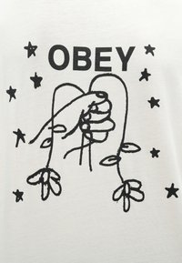 Wit T-shirt met een zwarte afbeelding van een hand die bloemen vasthoudt, met de tekst "OBEY" erboven en verspreide sterren rondom het ontwerp.