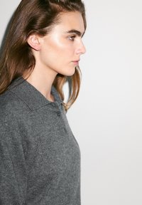 Šedý vlněný polo shirt s klasickým límečkem, s knoflíkovou légou a měkkou texturou, nošený osobou v profilu na neutrálním pozadí.