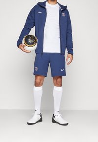 Tenue sportive Nike comprenant une veste bleu zippée, un t-shirt blanc, un short bleu, des chaussettes blanches et des chaussures de sport noires et blanches, tenant un ballon de football.