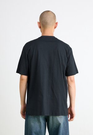 Osoba z ogoloną głową, ubrana w jednolity czarny t-shirt z krótkim rękawem i niebieskie dżinsy, widziana od tyłu na białym tle.