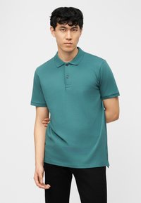 Jeune homme aux cheveux noirs courts portant un polo à manches courtes bleu sarcelle et un pantalon noir, debout devant un fond blanc.