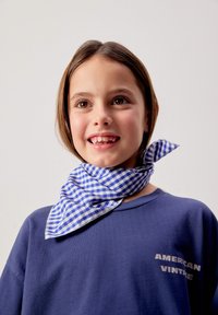 Young girl smiling, wearing a blue checkered neck scarf and a navy sweatshirt with "AMERICAN VINTAGE" text on a plain light background.
