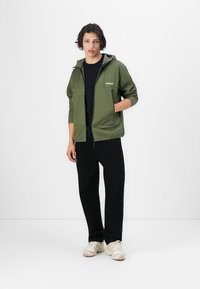Oliven grøn zip-up hættetrøje med sorte detaljer, der har en frontlomme og en hætte, båret over en sort skjorte og sorte bukser, parret med beige sneakers.