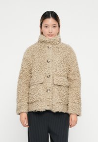Giacca in pelliccia sintetica beige con colletto alto, caratterizzata da grandi bottoni e due tasche frontali. Materiale texturizzato dall'aspetto accogliente.