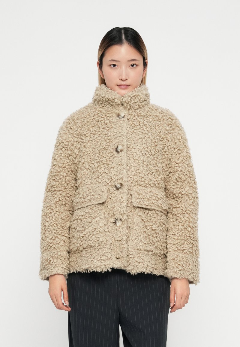 Beige neplederen jas met hoge kraag, voorzien van grote knopen en twee frontzakken. Gelaagd materiaal met een gezellige uitstraling.