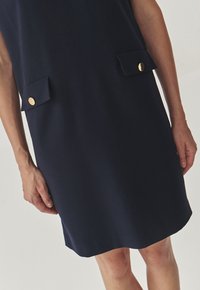 Robe sans manches bleu marine en tissu lisse, dotée de deux poches avant avec des boutons dorés. L'ourlet est droit et se situe juste au-dessus du genou.