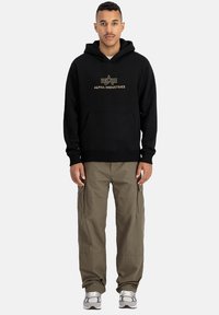 Hanorac negru cu logo-ul auriu "Alpha Industries", buzunar kangaroo, purtat cu pantaloni cargo oliv și adidași gri.