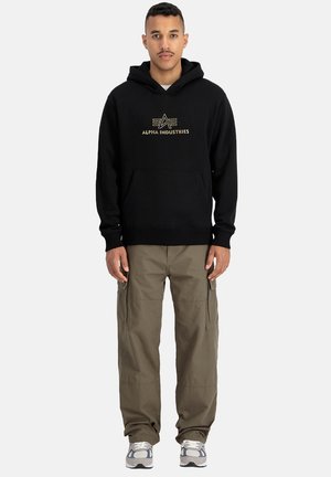 Zwarte hoodie met goudkleurig "Alpha Industries" logo, kangoeroezak, gedragen met olijfgroene cargobroek en grijze sneakers.