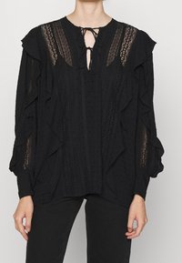 Blusa nera con volant, maniche lunghe, realizzata in tessuto texturizzato, con pannelli in pizzo trasparente e dettaglio alla scollatura con la possibilità di fare un nodo.