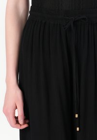 Black textured skirt with an elastic waistband, featuring a drawstring tie and gold-tone accents. Close-up of fabric and fastening details.