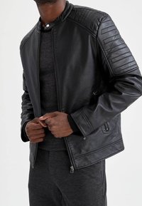 Veste en cuir noir avec des détails matelassés sur les épaules, des poches zippées et un col pressionné. Portée sur une couche en tricot texturé gris.