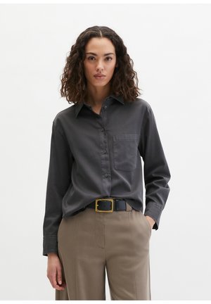 Femme aux cheveux bouclés portant une chemise gris foncé boutonnée rentrée dans un pantalon beige avec une ceinture noire, debout avec une main dans la poche.