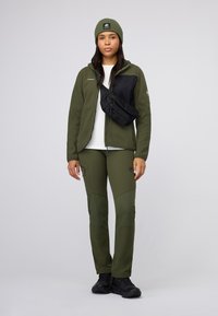 Olive green fleece jacket with black panel, olive pants, and black crossbody bag. Model wears black shoes and a matching olive beanie.