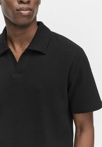 Svart polo skjorta tillverkad av texturerad tyg, med kort krage och halvknäppning. En enkel design med en figursydd silhuett.