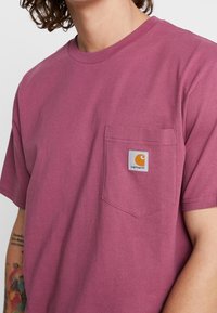 Kurzärmliges T-Shirt mit Rundhalsausschnitt in tiefem Mauve, aus Baumwolle, mit einer Brusttasche, die ein Carhartt-Logo-Label trägt.