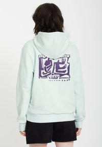 Felpa verde menta chiaro con una grande grafica viola sulla schiena, caratterizzata da figure astratte, polsini a costine e cappuccio con lacci.