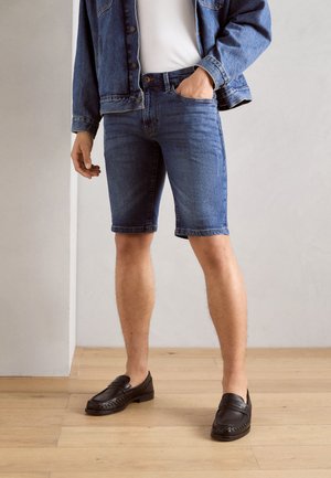 Shorts di jeans blu, con un taglio classico e orlo appena sopra il ginocchio. Abbinati a mocassini scuri e una t-shirt bianca sotto una giacca di jeans.