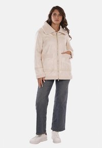 Giacca in pelliccia sintetica beige con colletto ampio, due tasche frontali e zip completa. Abbinata a jeans azzurri e sneakers bianche.
