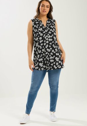 Kvinde iført sort ærmeløs top med hvidt tusindfrydprint, blå tætsiddende jeans og hvide sneakers, stående med hænderne i lommerne.