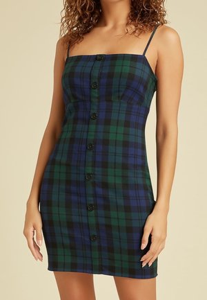 Kobieta ubrana w bezrękawnikową, rozpinaną mini sukienkę w zielono-niebieską kratę z cienkimi ramiączkami.