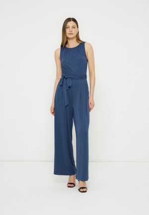 Kvinne iført ermeløs marineblå jumpsuit med vide ben og midjebånd, og svarte åpne hæler, stående mot en ensfarget hvit bakgrunn.