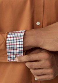 Hele pruun särk, millel on punased, valged ja sinised ruudulised varrukaotsad. Särgil on nööbist alla keeratav krae ja esiküljel tooni järgi nööbid.