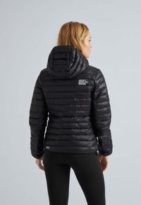 Svart puffjacka med huva, med kviltade linjer och logodetaljer på ryggen och fållen, tillverkad av ett lätt, glänsande material.
