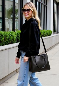 Borsa a tracolla in pelle nera con maniglie doppie e tracolla, forma rettangolare, texture liscia, abbinata a jeans a gamba larga azzurri e una felpa nera.