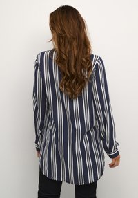 Camisa de manga larga azul marino con rayas verticales blancas, de tejido suave y con un dobladillo curvado. Presenta puños abotonados y un ajuste relajado.