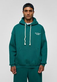 Teal hoodie met een voorzak, roomkleurige trekkoorden en geborduurd logo op de borst. Soepele stofstructuur.
